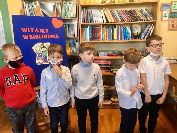 Pięciu chłopców na tle tablicy z napisem Witamy w bibliotece. Z tyłu regały z książkami