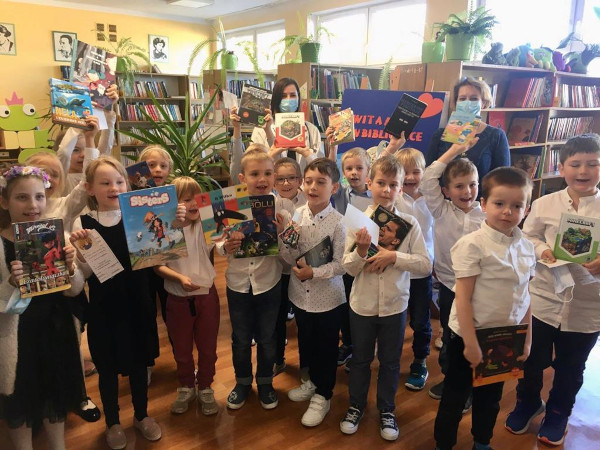 Uczniowie w bibliotece. W dłoniach trzymają książki. Z tyłu stoją panie bibliotekarki.