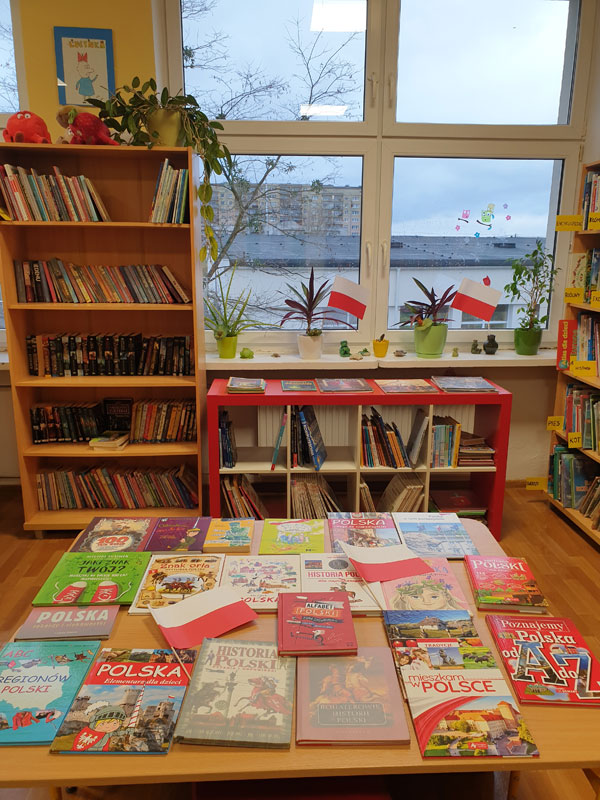Na podłodze leżą książki związane z Polską.W tle regały z książkami i okno, na parapecie, którego są kwiaty. Do doniczek wsunięte są dwie flagi Polski.