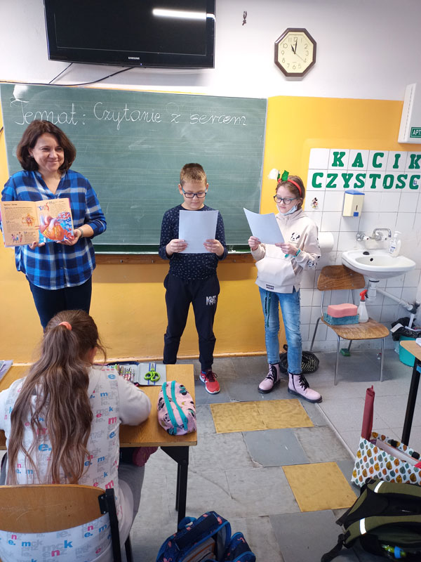 Nauczyciel bibliotekarz z książką, dziewczynka i chłopiec z kartkami w dłoniach, w tle tablica szkolna.