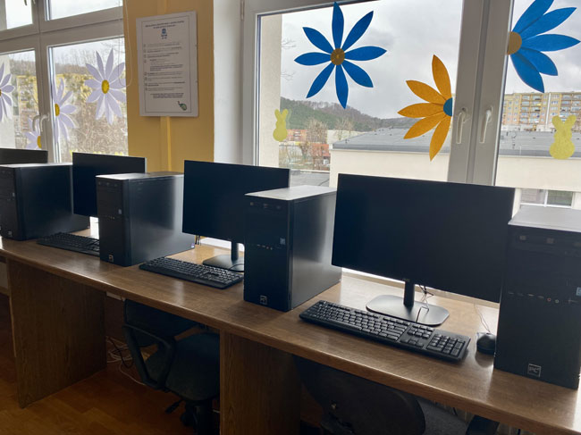 Komputery na tle okna, na oknach kolorowe kwiaty.