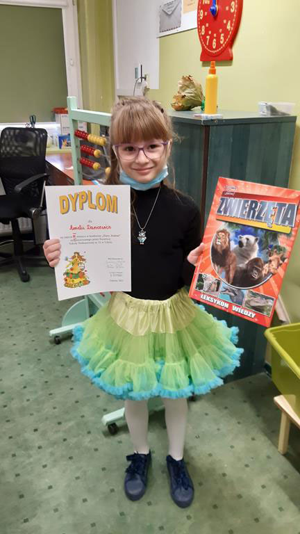 Stojąca dziewczynka trzyma w prawej dłoni dyplom, w drugiej książkę 'Zwierzęta'.