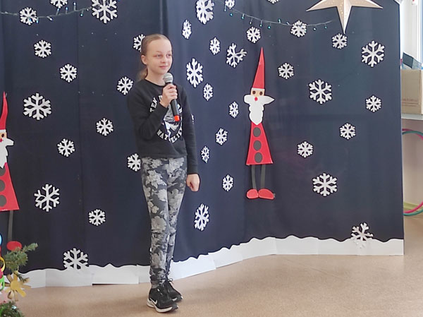 Dziewczynka z mikrofonem, granatowe tło, białe gwiazdki, dwa czerwone mikołaje z papieru.