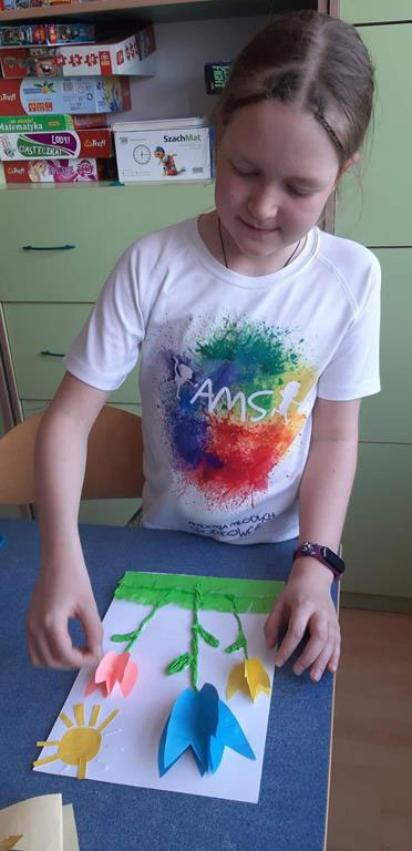 Dziewczynka, na stole biała kartka z naklejonymi kwiatami, trawą, słońcem.